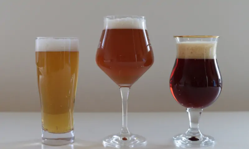 Drie glazen bier met schuim op een witte tafel: blond, amberkleurig en donker bier tegen een grijze achtergrond.