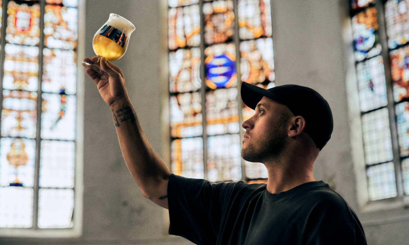 Saïd Kinos houdt een Duvel designerglas met blond bier omhoog voor kleurrijke glas-in-loodramen.