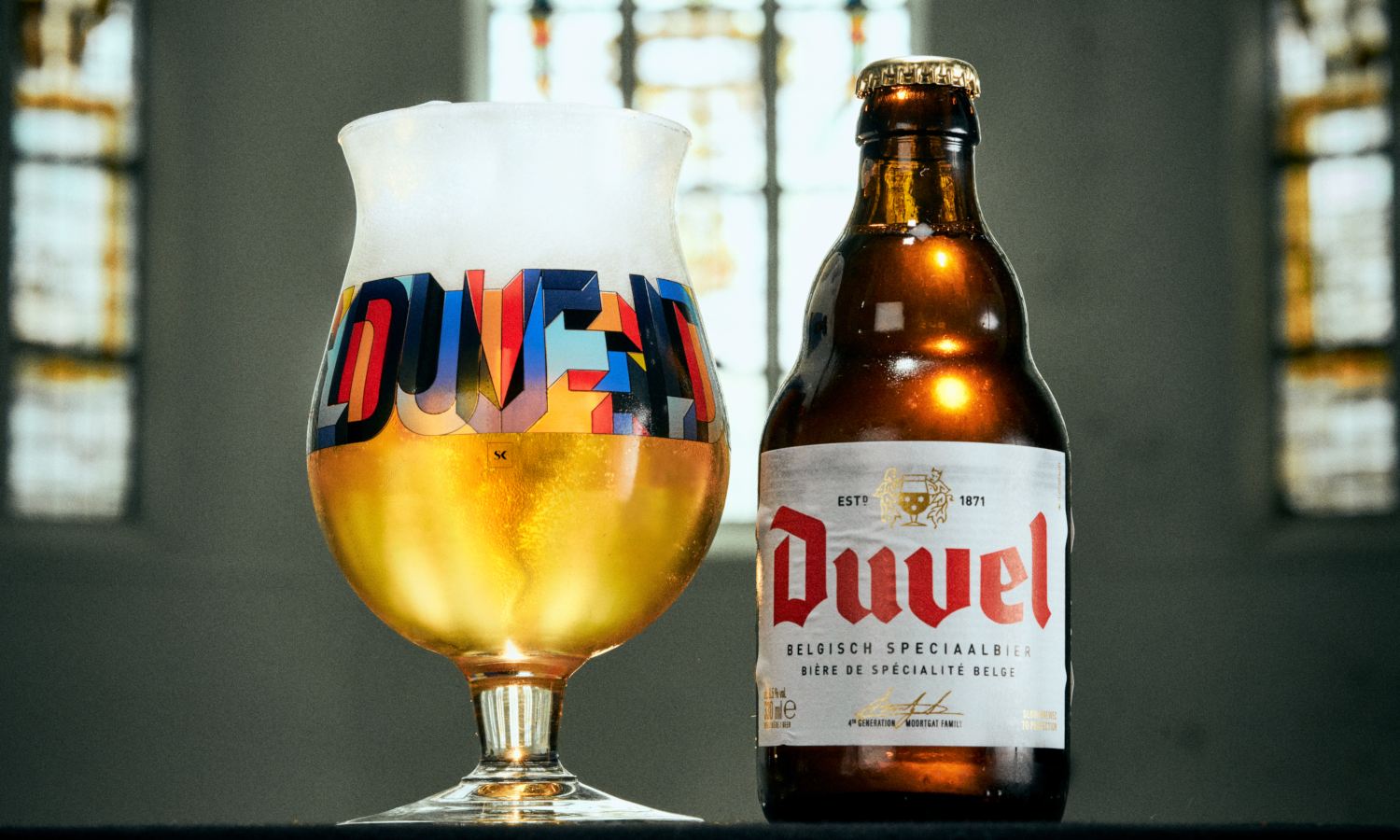 Duvel designerglas met schuimkraag en kleurrijk logo naast een Duvel fles voor onscherpe ramen.