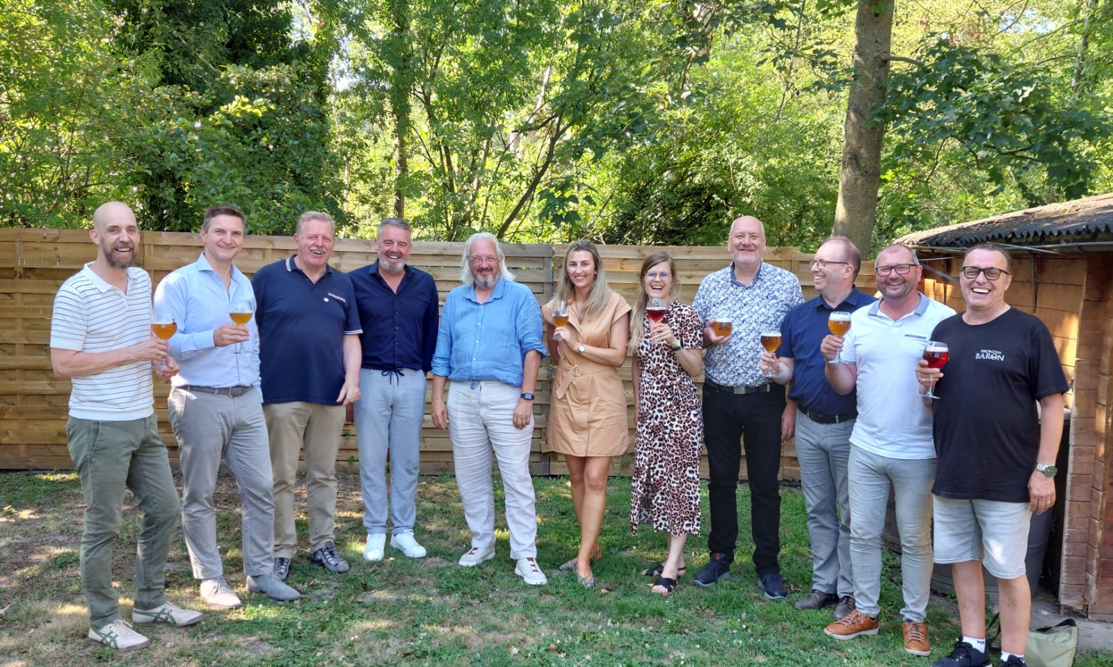 DKM Brewery werknemers proosten met bierglazen in een groene tuin bij een houten omheining.