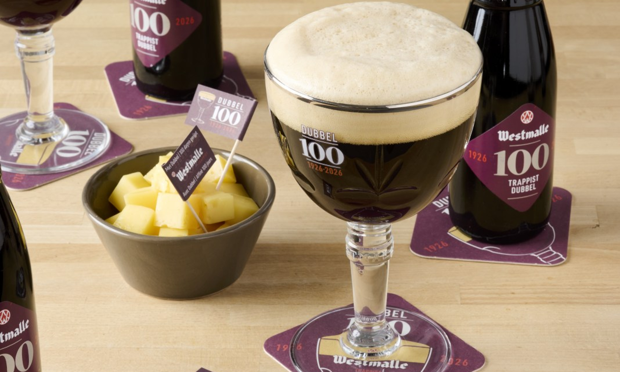 Glas Westmalle Dubbel 100 met schuimkraag, flesjes en kaasblokjes op een houten tafel.