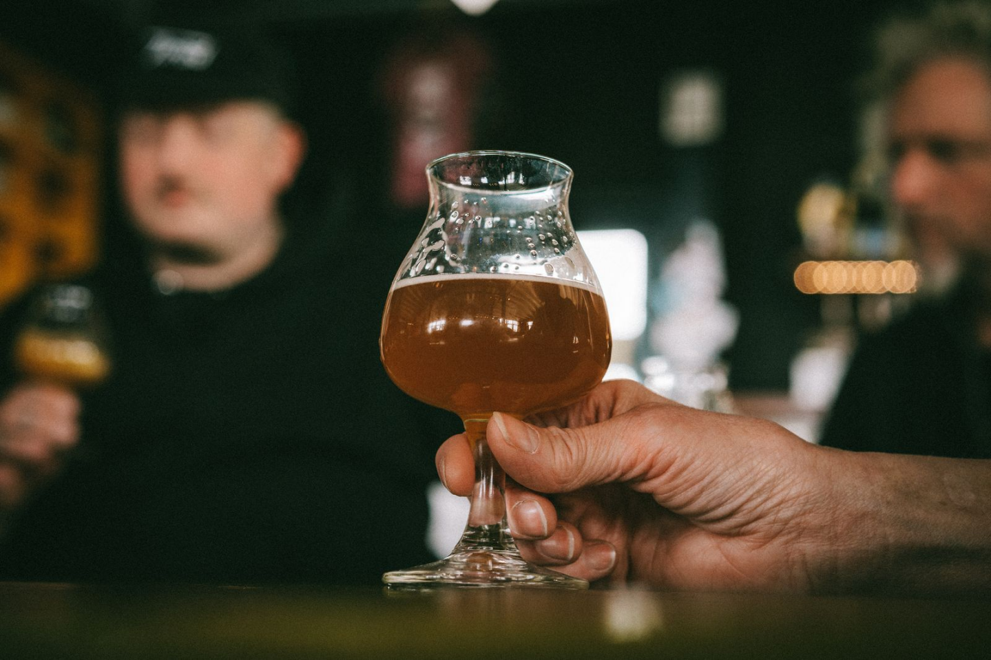 Hand houdt een tulpglas met amberkleurig bier vast in een donkere bar.