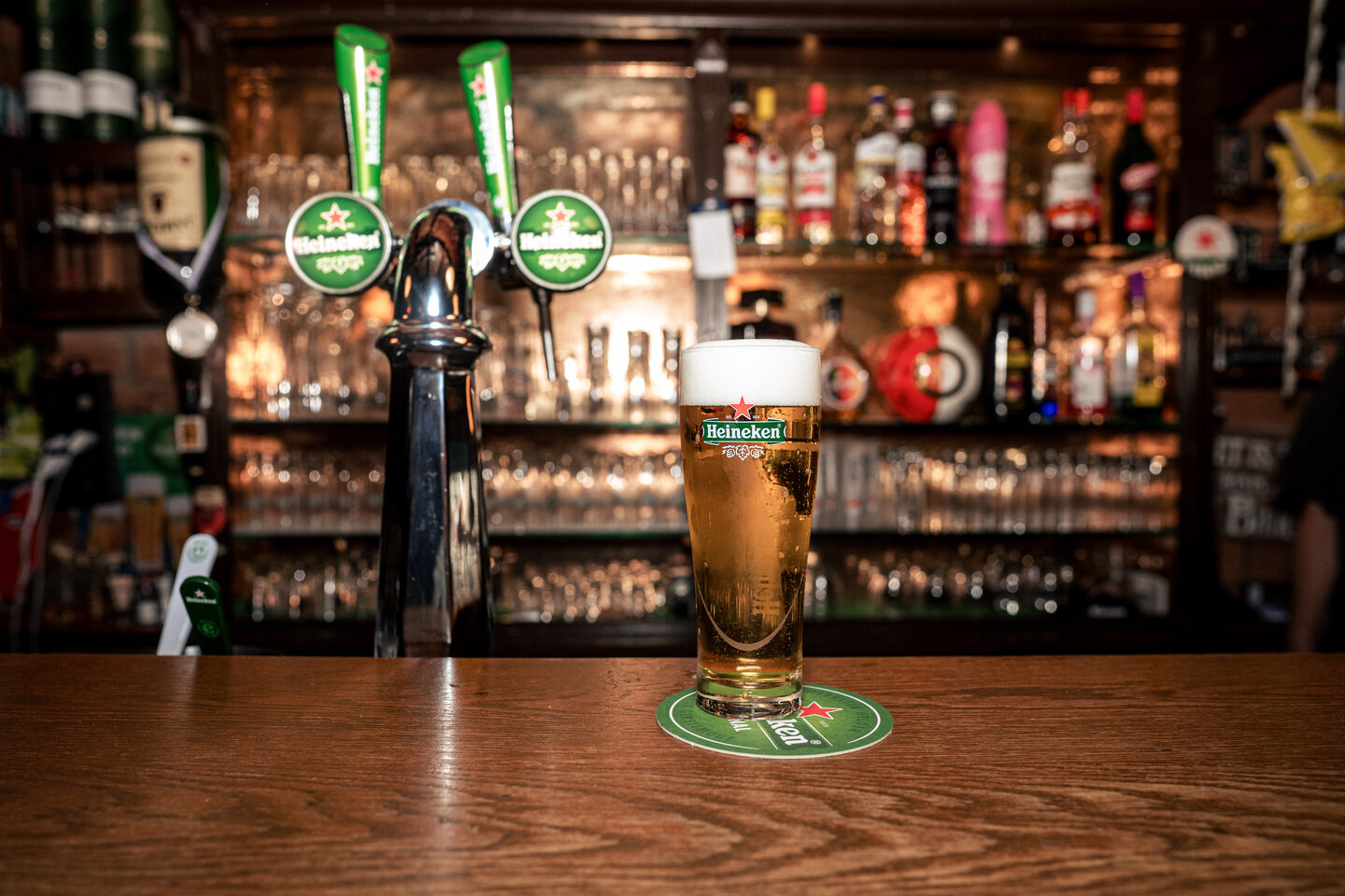 Glas vers getapte Heineken met stevige schuimkraag naast een tapzuil op een houten bar.