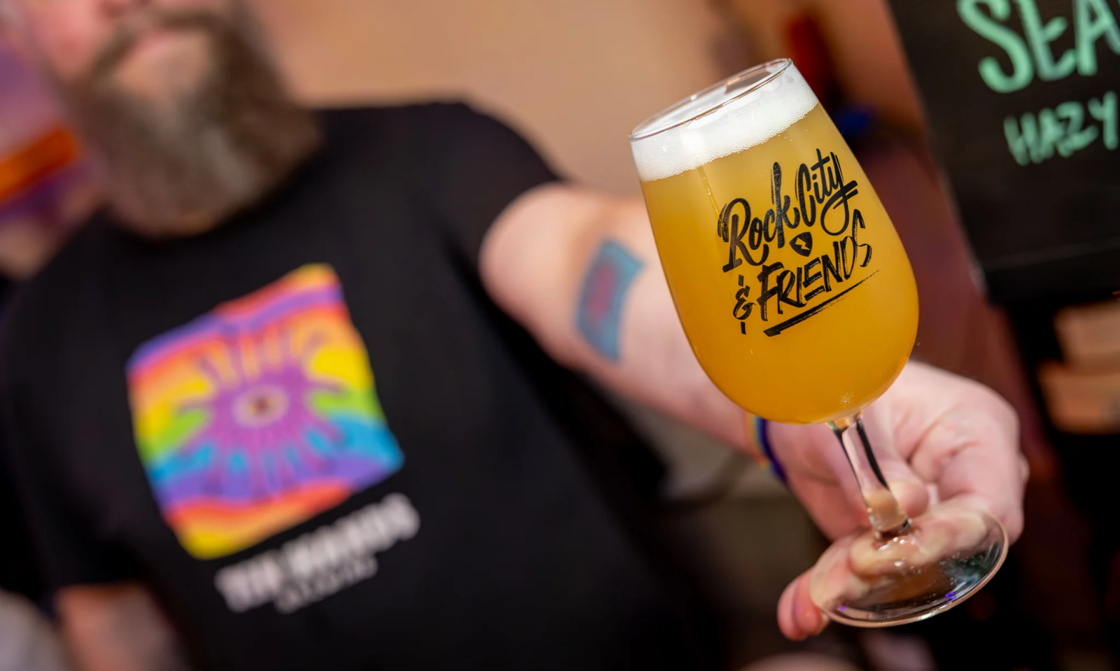 Hand houdt een glas troebel geel bier met Rock City & Friends-logo, met onscherpe persoon op de achtergrond.