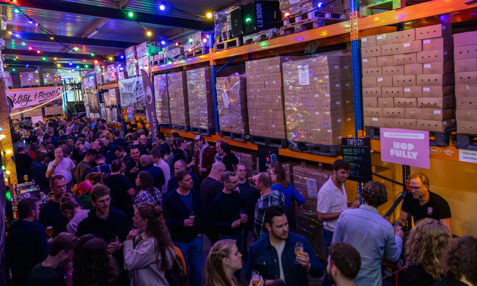 Drukke Rock City-bijeenkomst in een magazijn met bierdrinkende bezoekers en kleurrijke lichtslingers.
