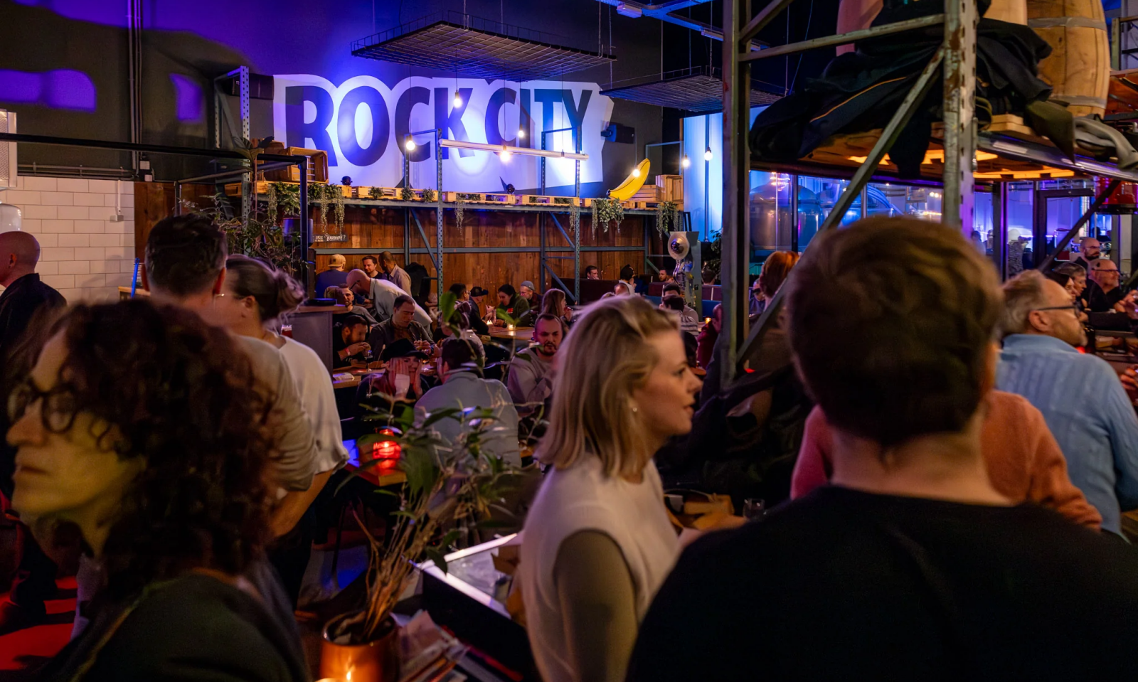 Drukke indoor horecaruimte met groot Rock City-logo, industriële inrichting en paarse verlichting.