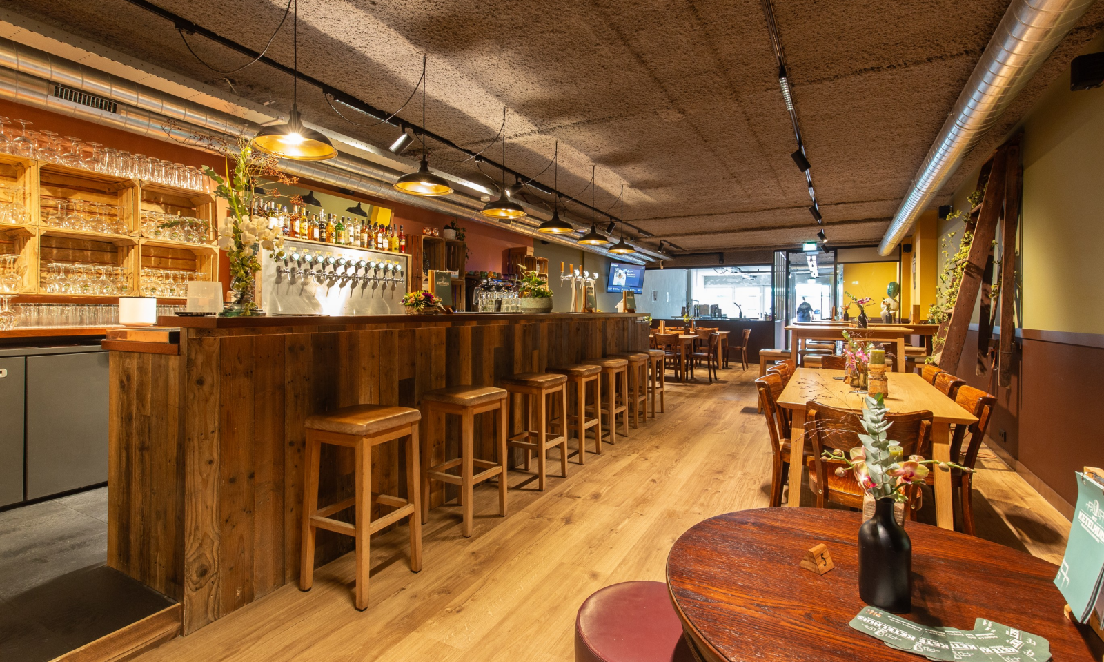 Warm interieur van biercafé met houten tafels, bar, tapkranen en industriële luchtbuizen.