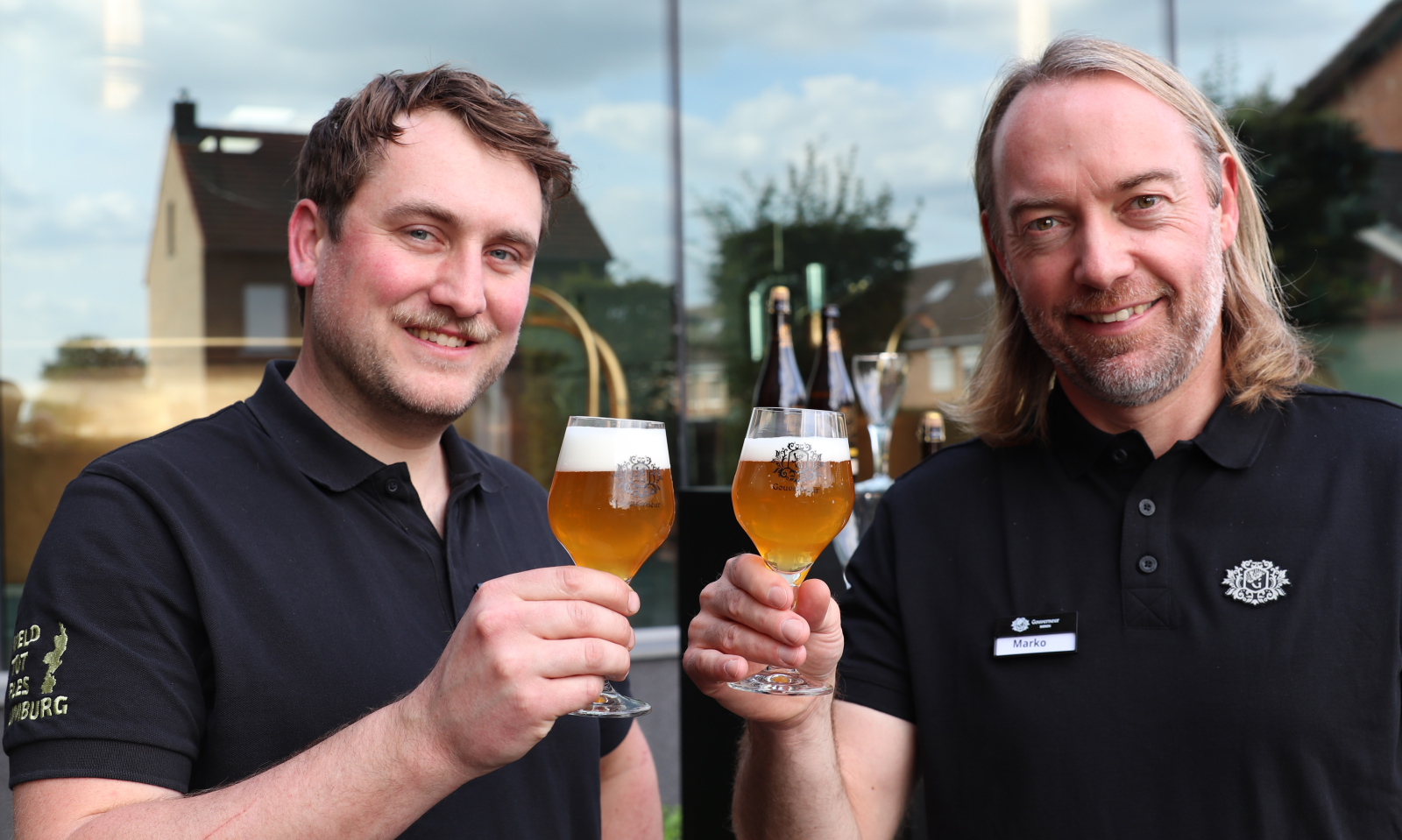 Willem Veldman en Marko Versteeg  in zwarte polo's proosten met glazen Exquise Blond Whisky Infused bier voor een raam.