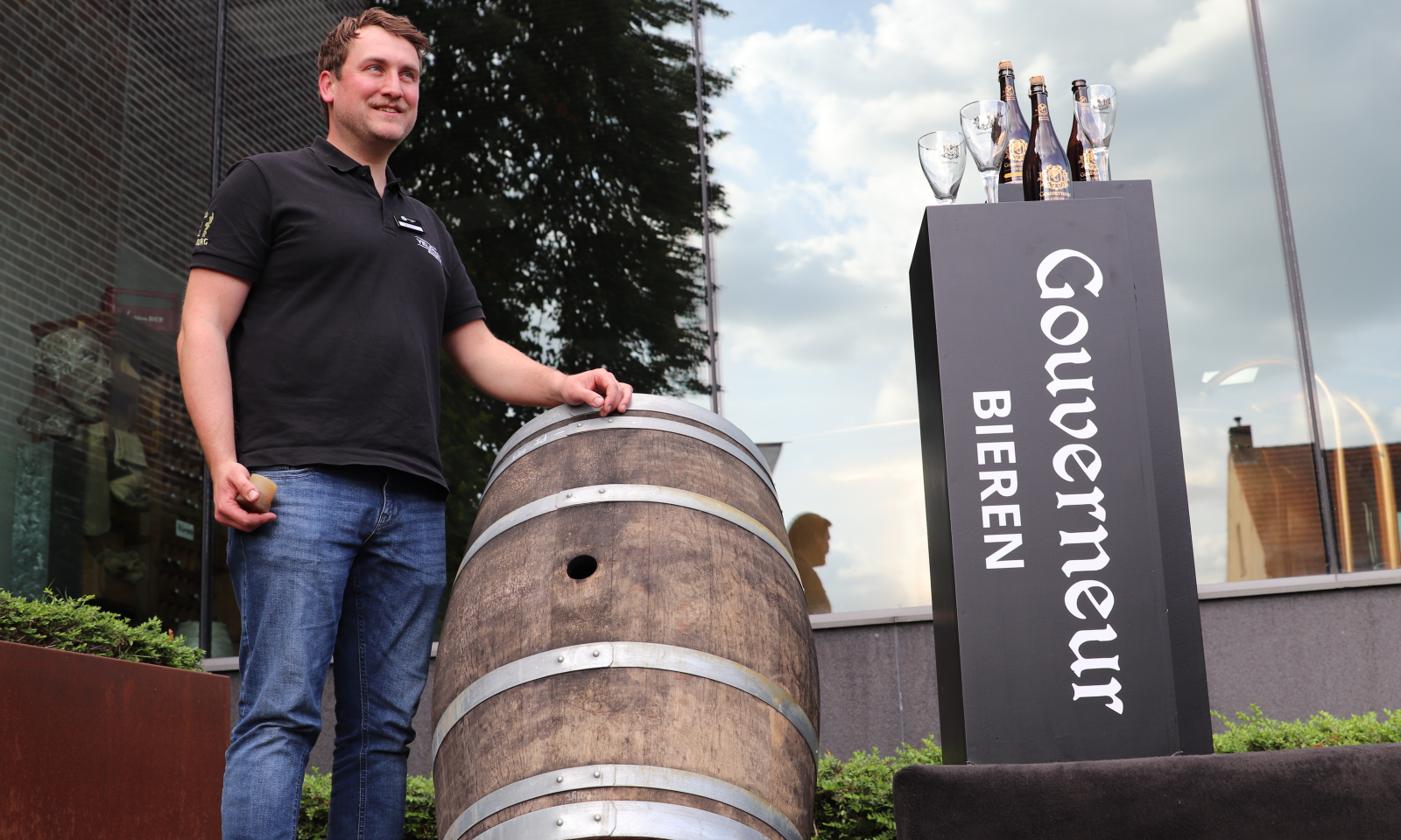 Willem Veldman naast houten vat en Gouverneur-bierdisplay met flessen en glazen voor een gebouw.