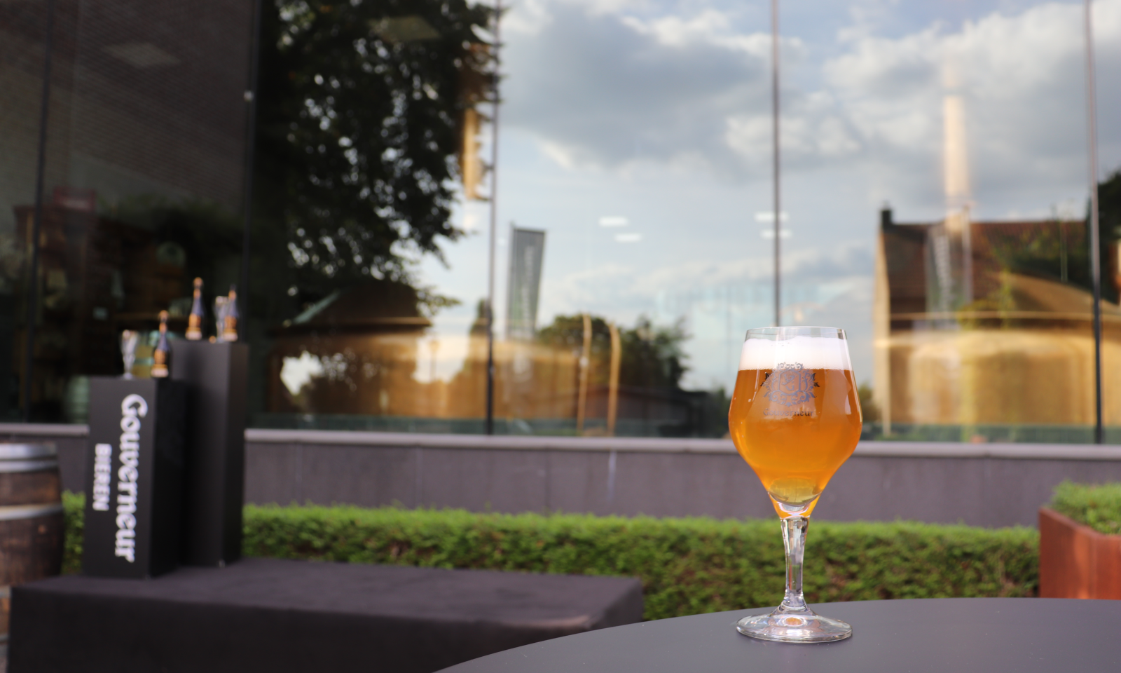 Glas amberkleurig Gouverneur-bier op een terras met promotiestand en flesjes op de achtergrond.
