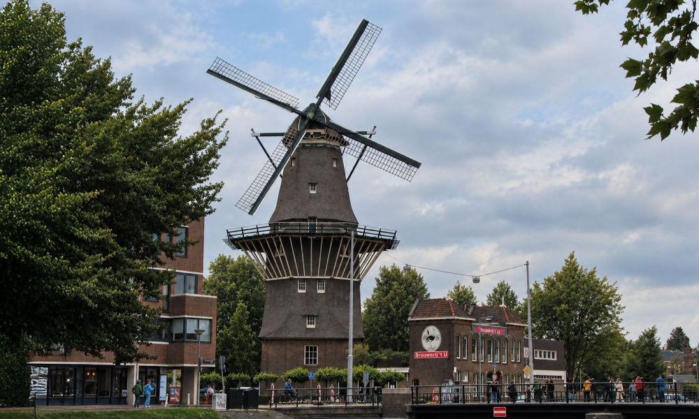 Molen De Gooyer bij Brouwerij ’t IJ in Amsterdam met bomen, brug en bewolkte lucht.