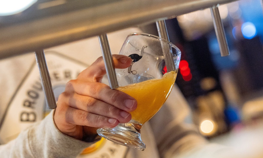 Hand houdt een proefglas met troebel geel bier onder een metalen tap tijdens een bierproeverij.