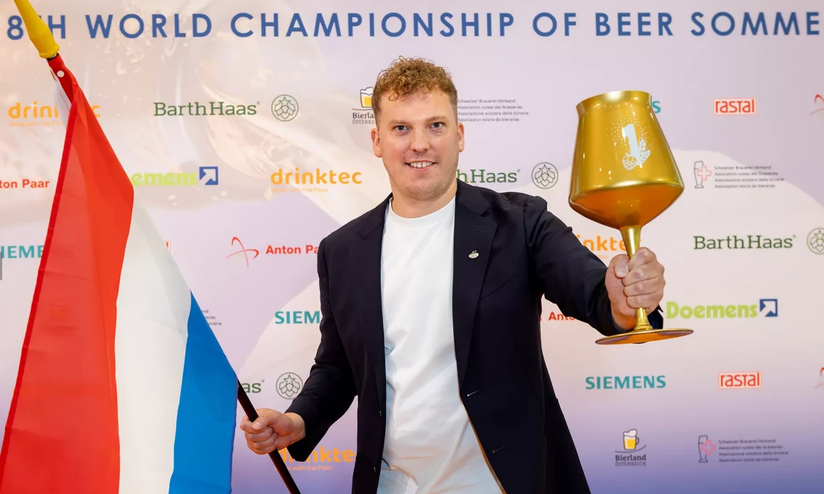 Léon Rodenburg met Nederlandse vlag en gouden bierglas voor sponsorwand van biersommelierkampioenschap.