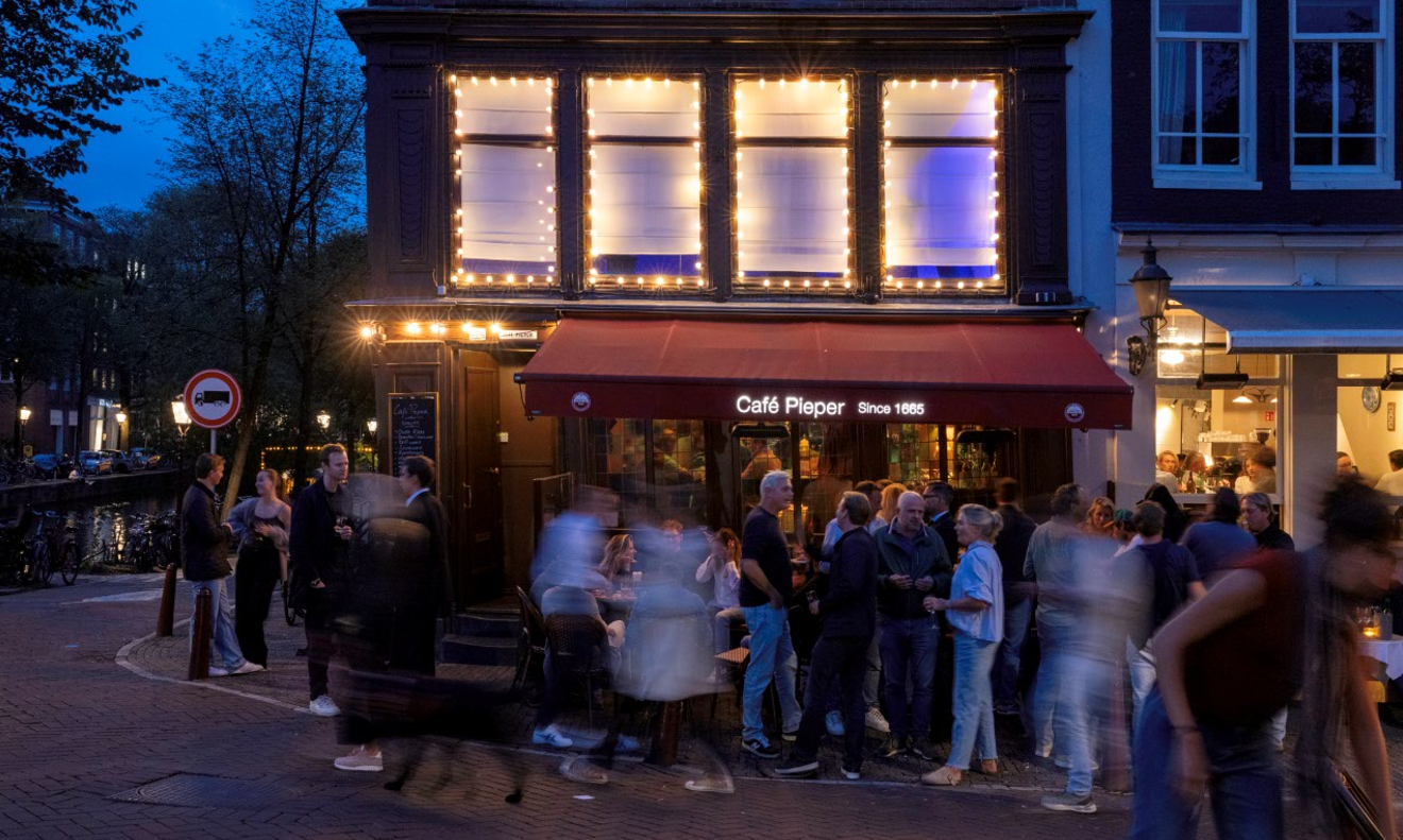 Druk terras voor Café Pieper in Amsterdam bij avond, met verlichte gevel en gasten op straat.