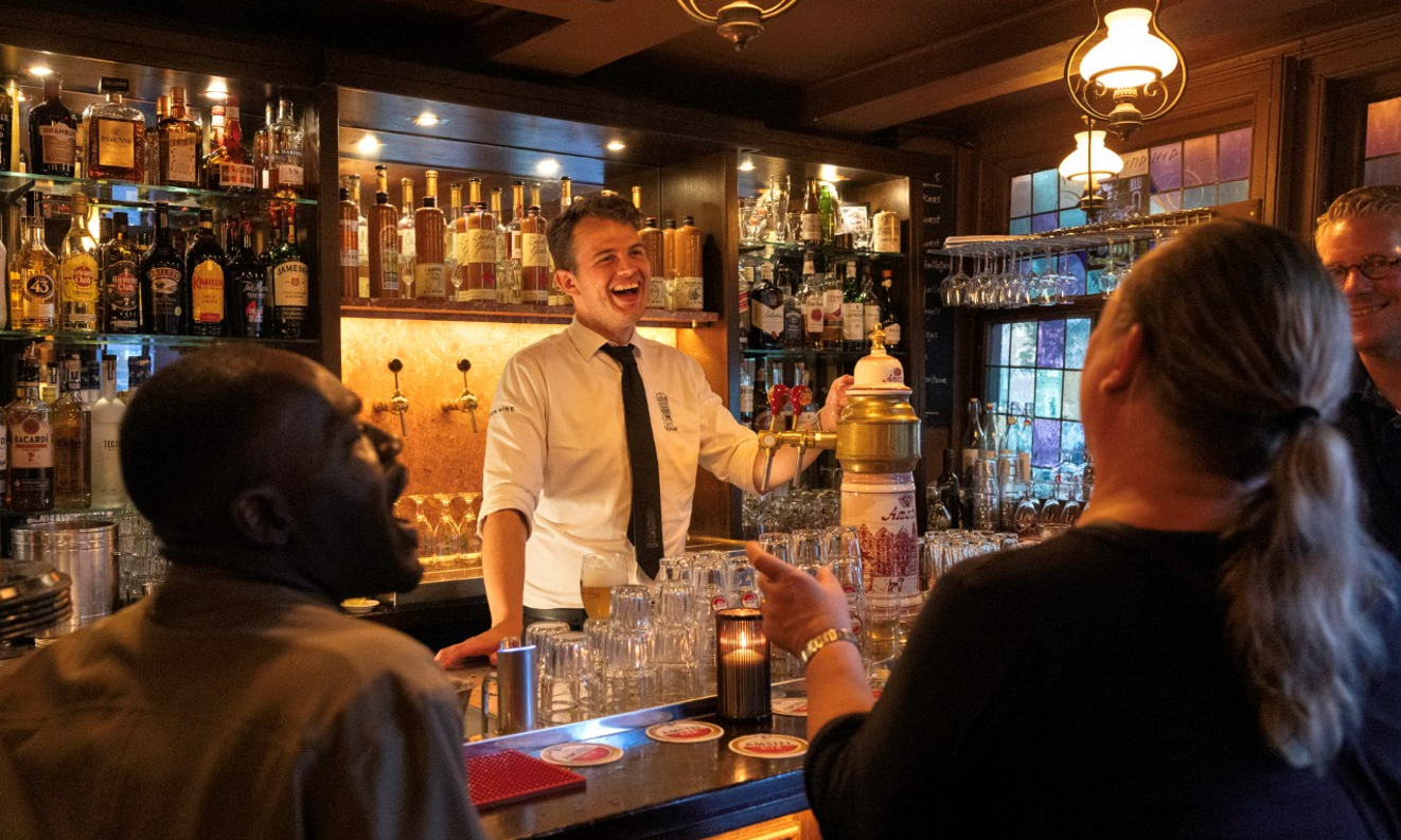 Lachende barman achter de bar van een bruine kroeg met gasten, drankflessen en warm licht.