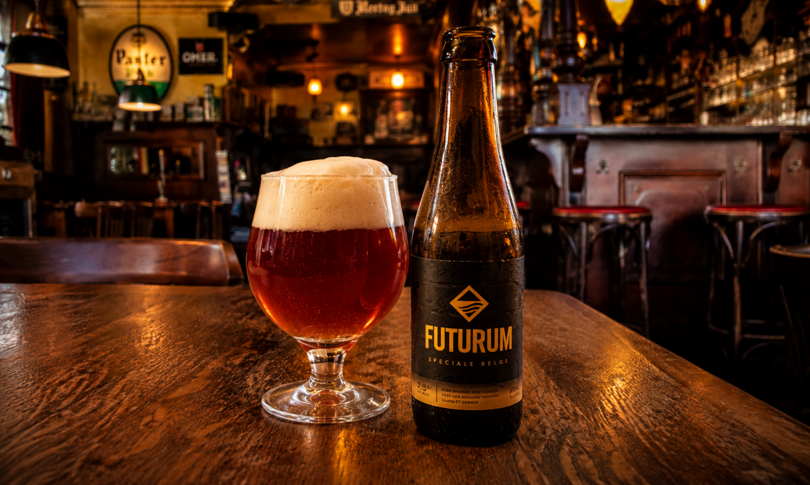 Glas amberkleurig bier met schuim naast een Futurum-fles op een houten tafel in een warme kroeg.