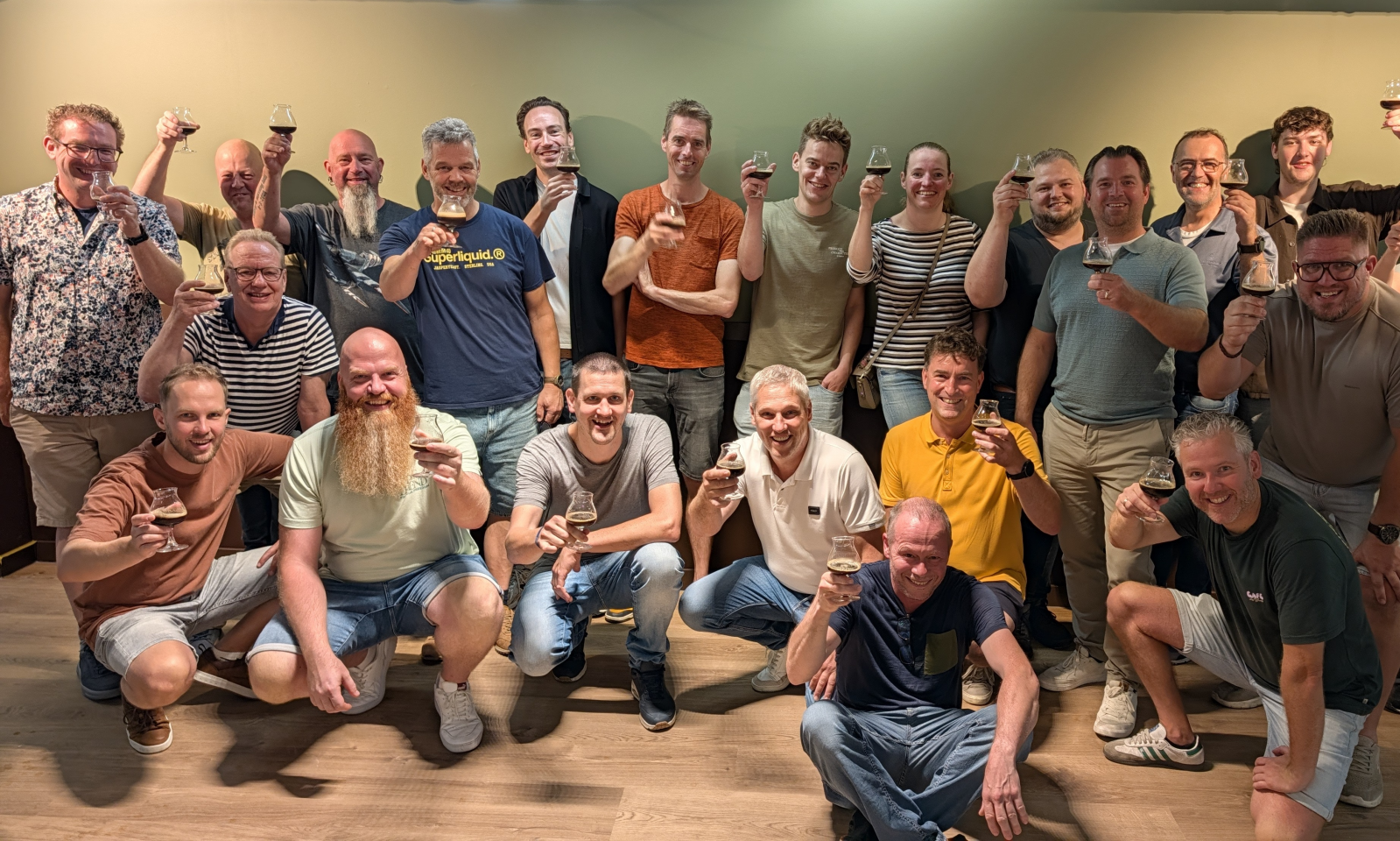 Groepsfoto van ongeveer twintig mensen die toosten met kleine bierglazen in een binnenruimte