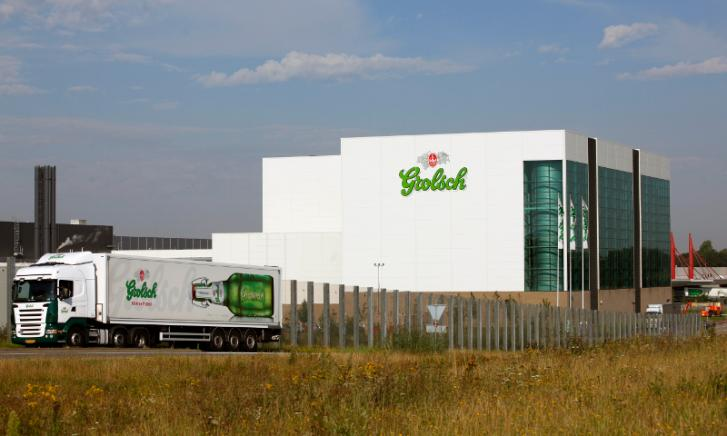 Grote witte fabriekshal met Grolsch-logo en vrachtwagen met bierreclame op een weg in het voorgrondgebied