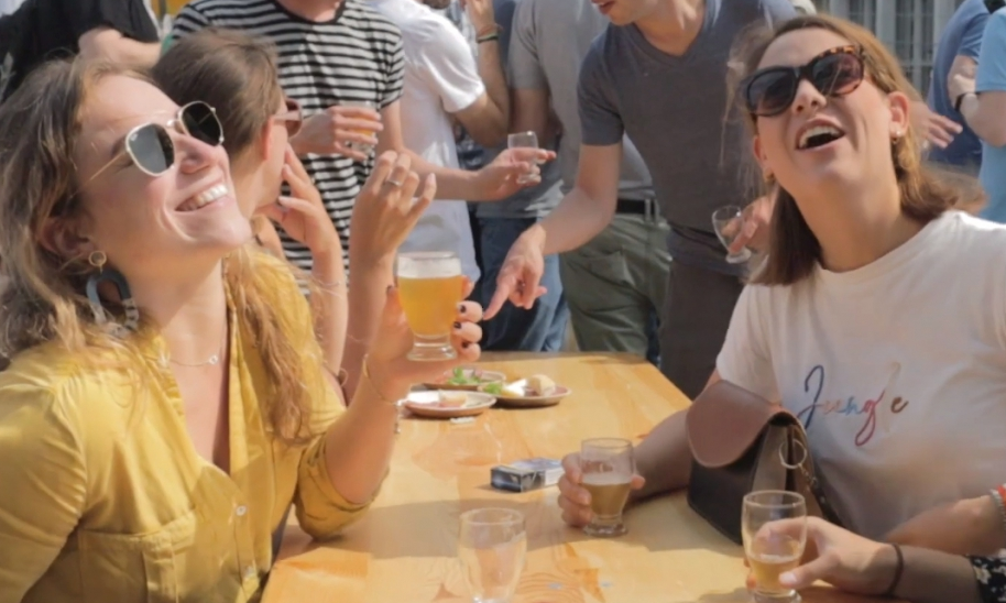 Drie lachende vrouwen aan een houten tafeltje met bier, zon en publiek op achtergrond