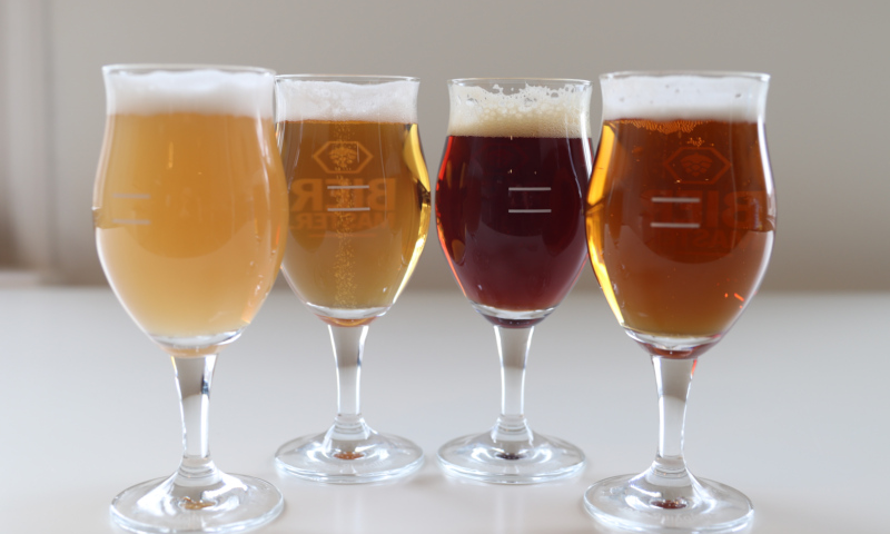 Vier verschillende biertjes in tulpglazen op een witte ondergrond, variërend in kleur en schuimkraag.
