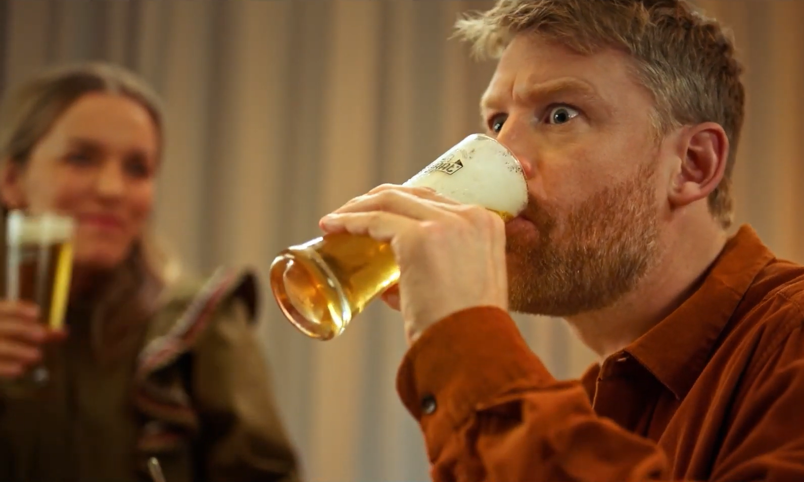Man drinkt bier uit een glas met schuim terwijl een andere persoon kijkt.