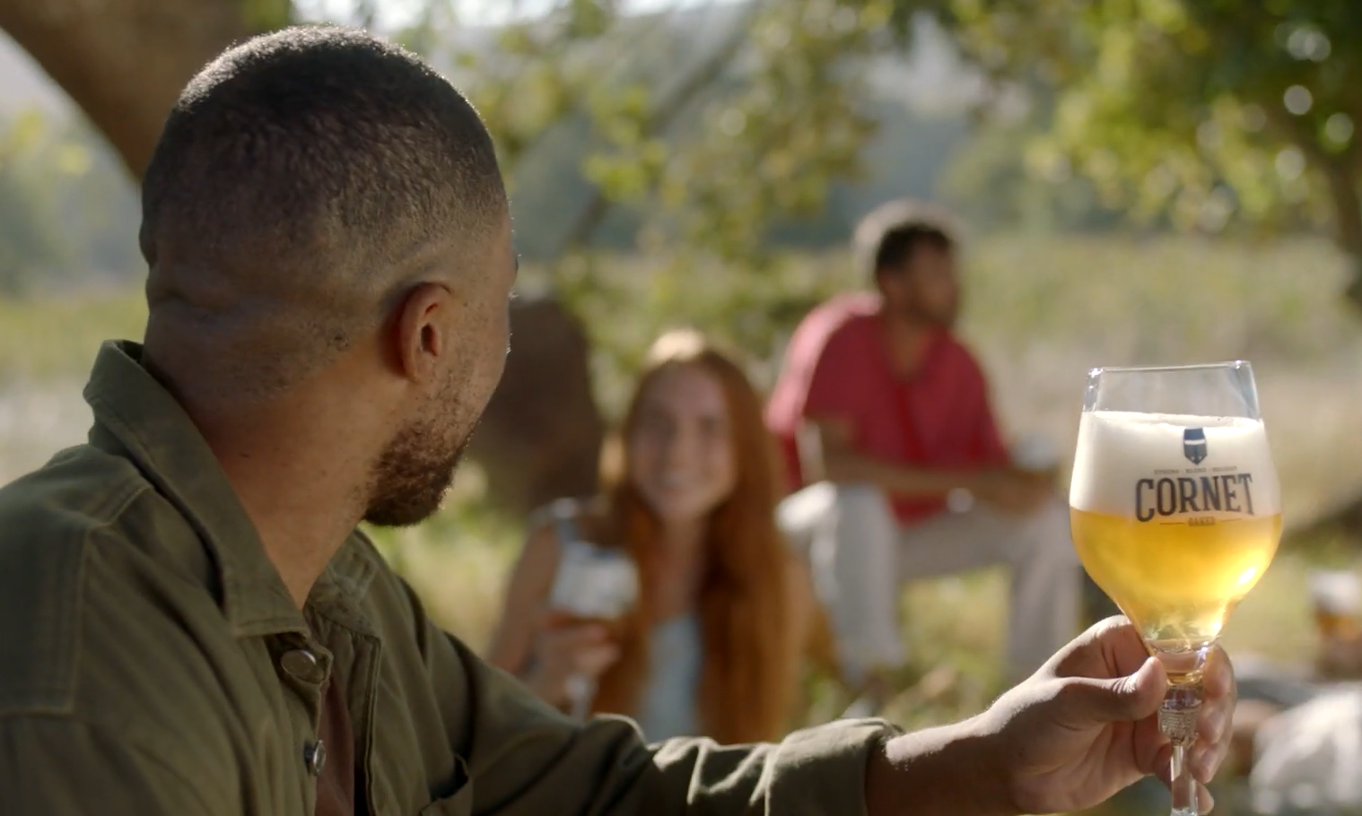 Man houdt een glas Cornet bier omhoog in een tuin met vrienden op de achtergrond.