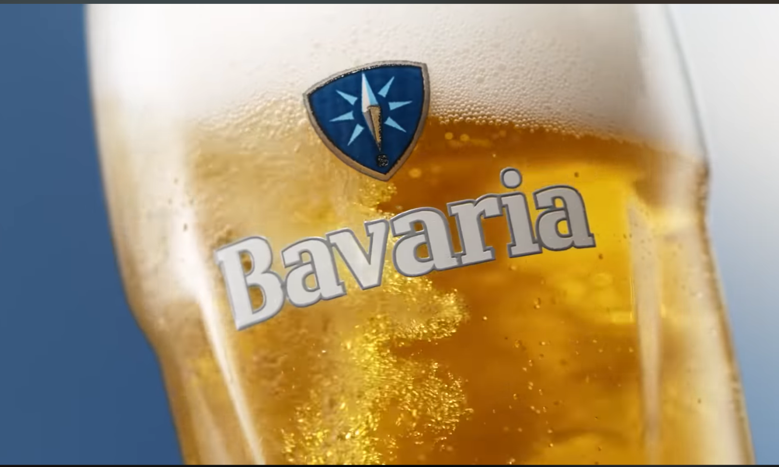 Glas met goudgele Bavaria 0.0% drank met schuimkraag op een blauwe achtergrond.