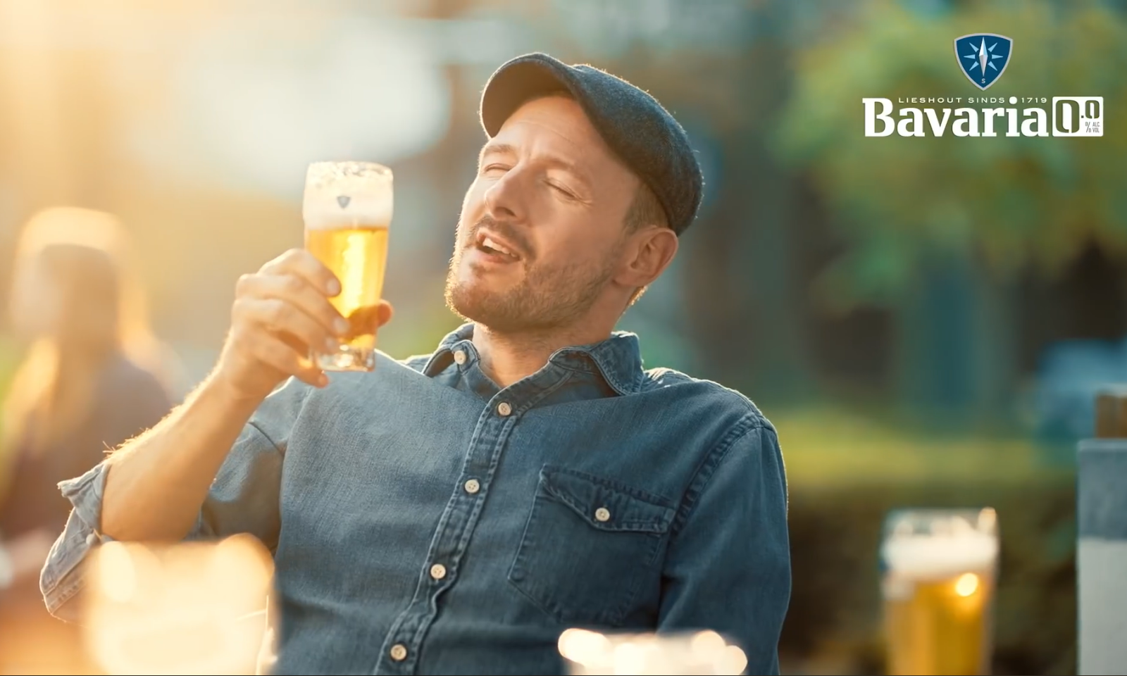 Man drinkt bier in de zon met Bavaria logo op de achtergrond.