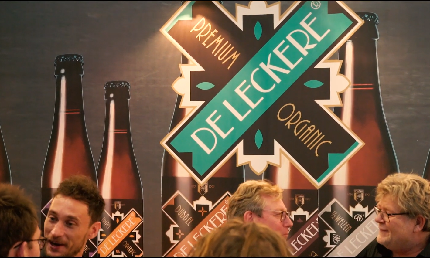 Groep mensen voor een reclamedisplay van Brouwerij de Leckere met bierflesjes en logo.