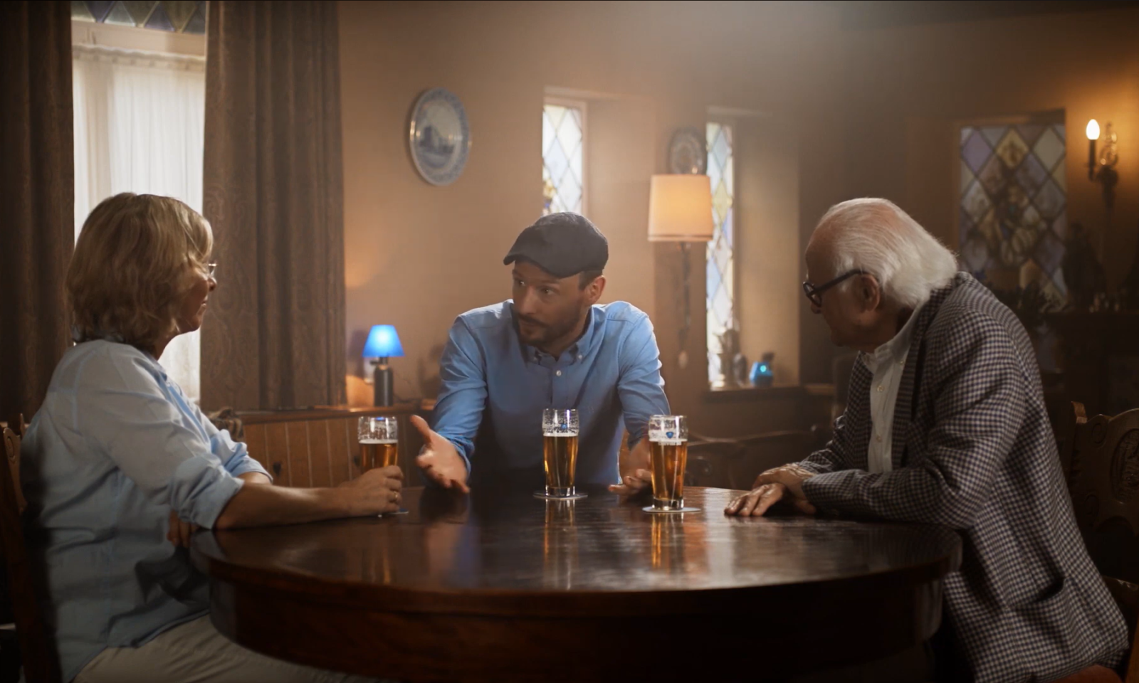Drie mensen aan een ronde houten tafel met bierglazen in een warme, sfeervolle kamer.