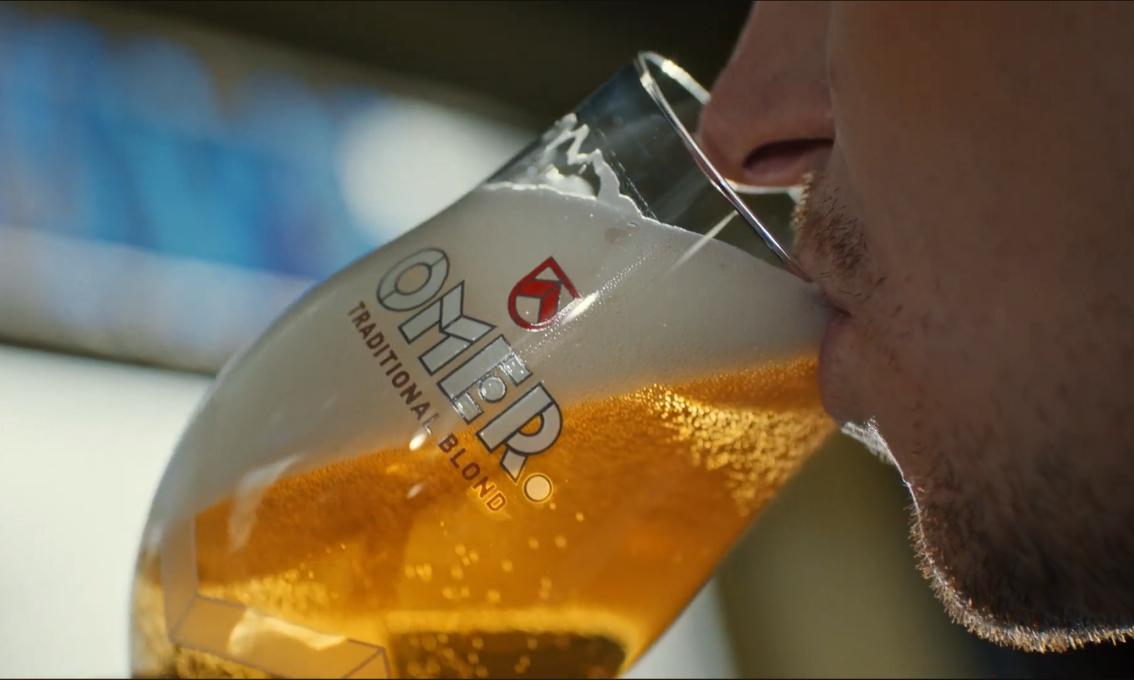 Close-up van een persoon die een glas Omer Traditional Blond bier drinkt.