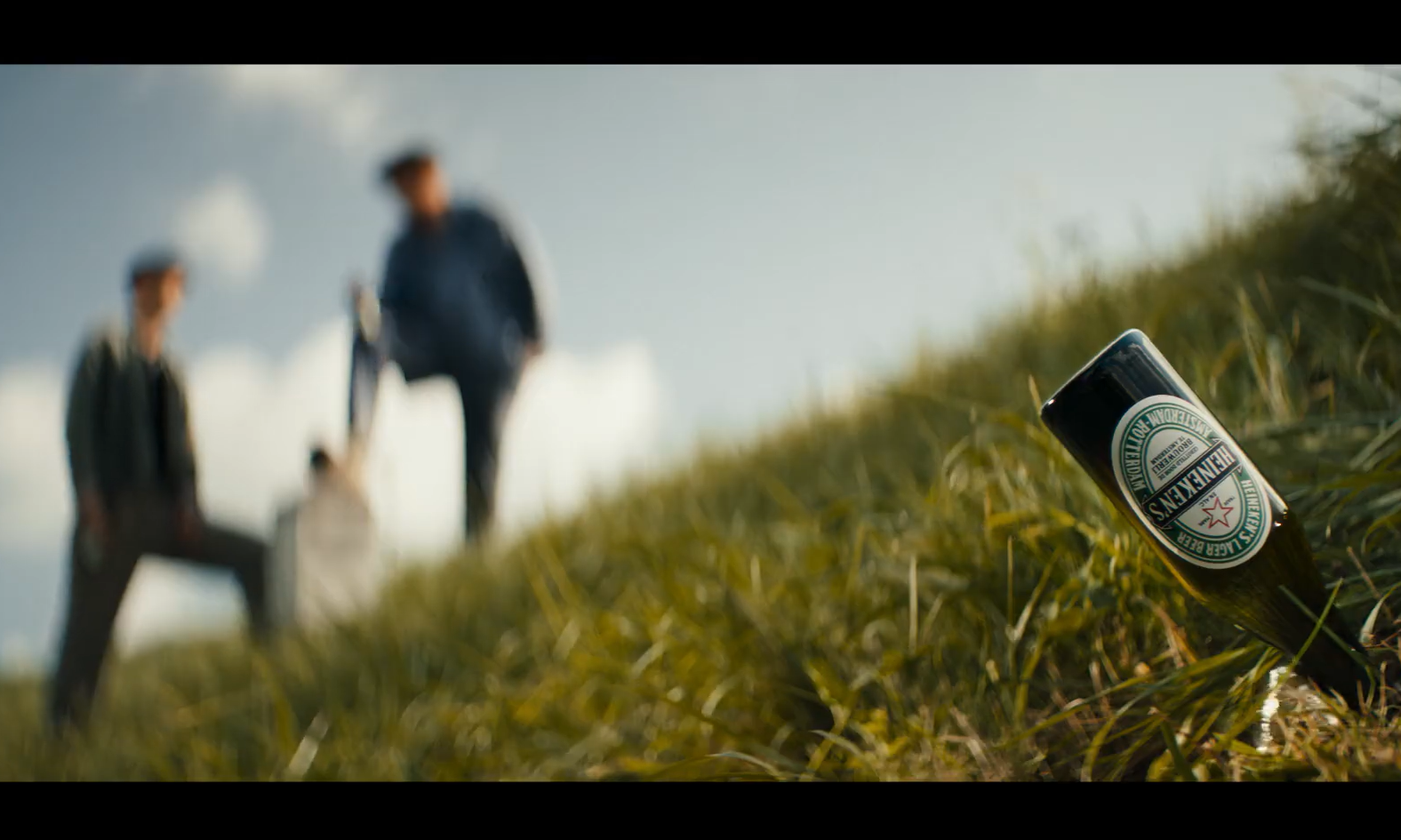 Een Heineken bierfles ligt in het gras met onscherpe mensen op de achtergrond.