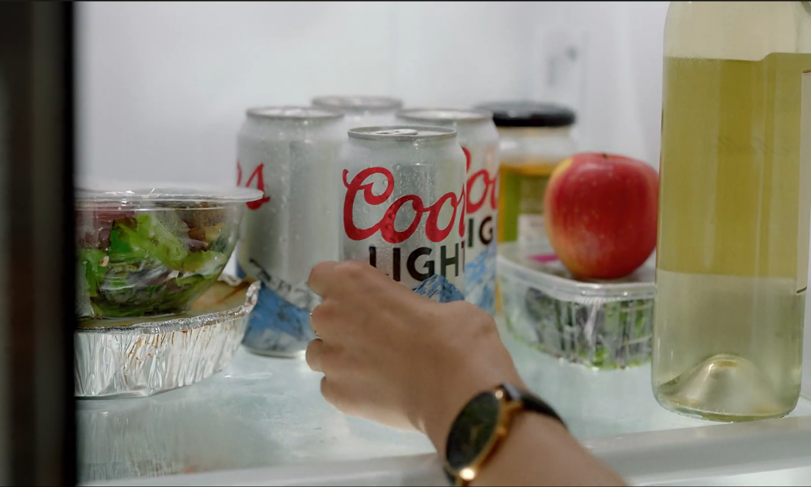Hand pakt een blik Coors Light bier uit een koelkast naast salade en appel.