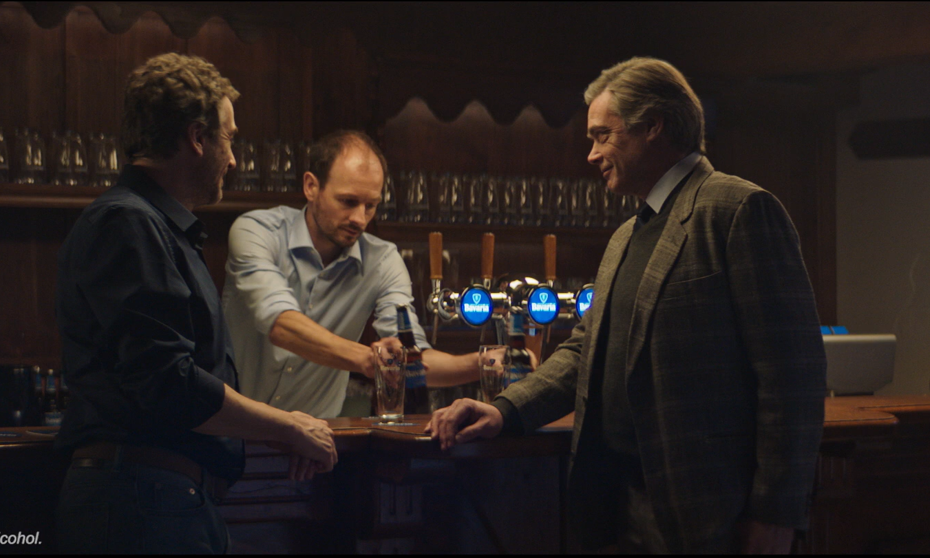 Drie mannen aan een bar, een barman schenkt een glas bier met Bavaria logo.