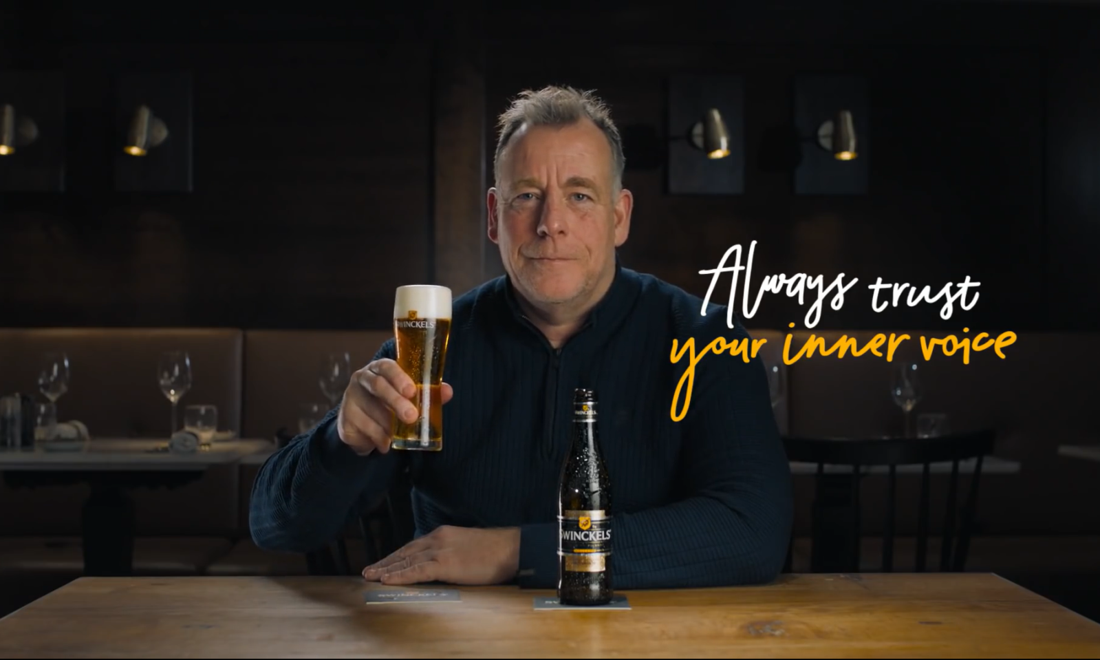 Man houdt een biertje omhoog in een restaurantomgeving, met de tekst 