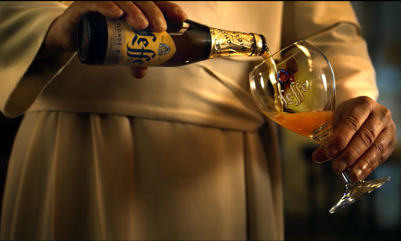Een persoon in een monnikspij schenkt Leffe bier in een glas met logo.