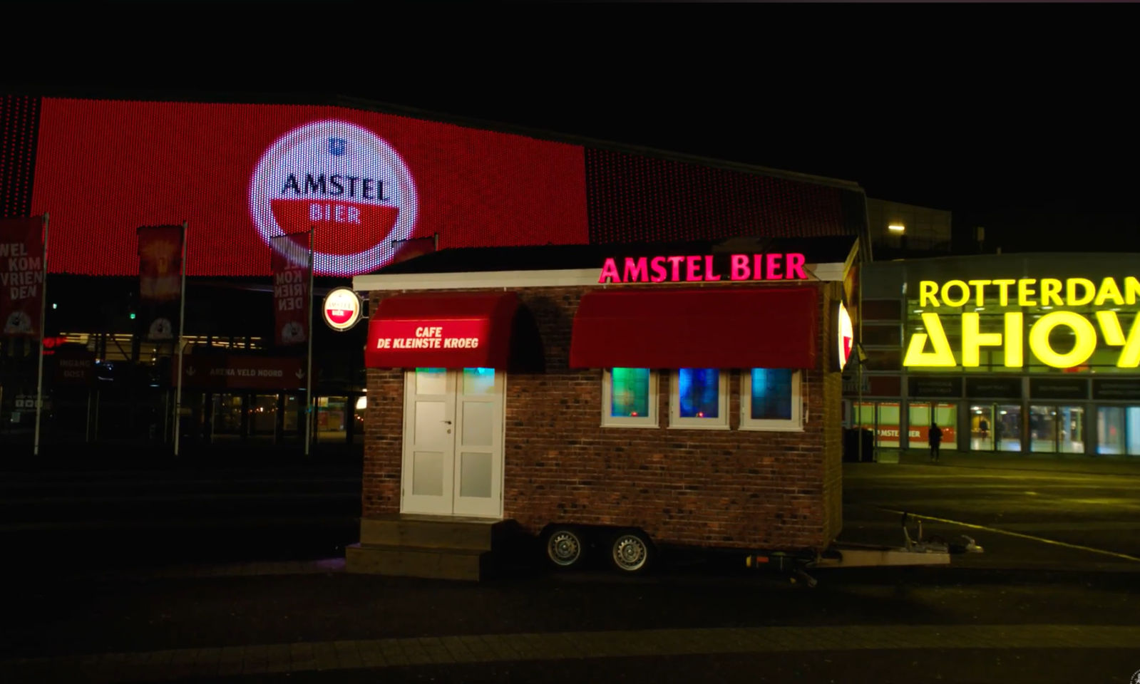 Klein café met rood Amstel-bierbord voor de verlichte Ahoy in Rotterdam.