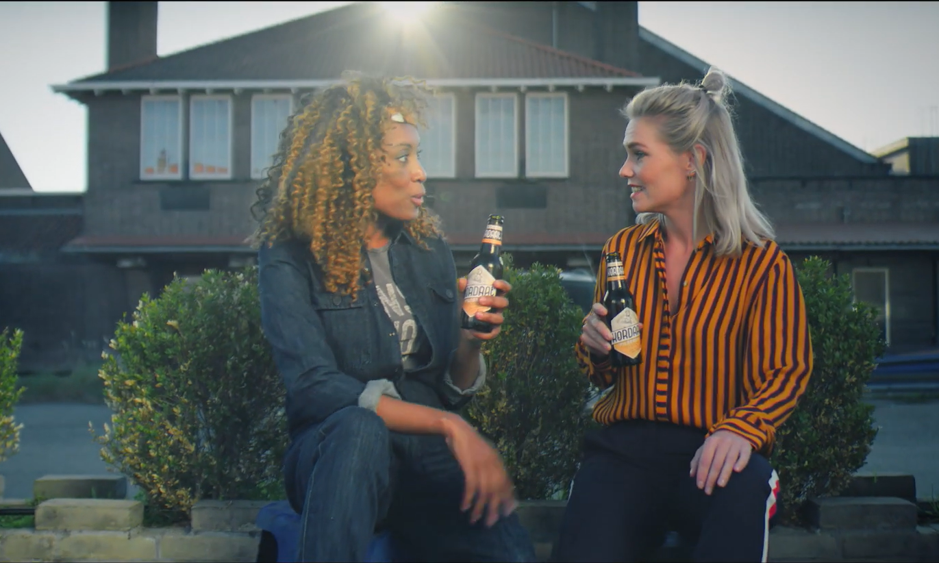 Twee vrouwen zitten op een muurtje, houden flesjes bier vast en praten met elkaar.
