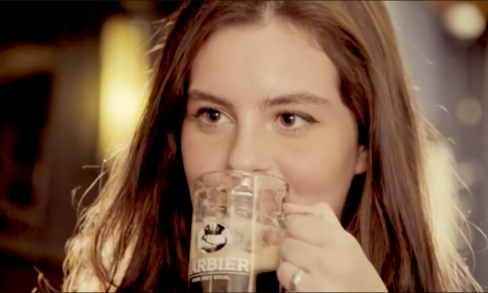 Vrouw drinkt een donker biertje uit een glas met Barbier logo.