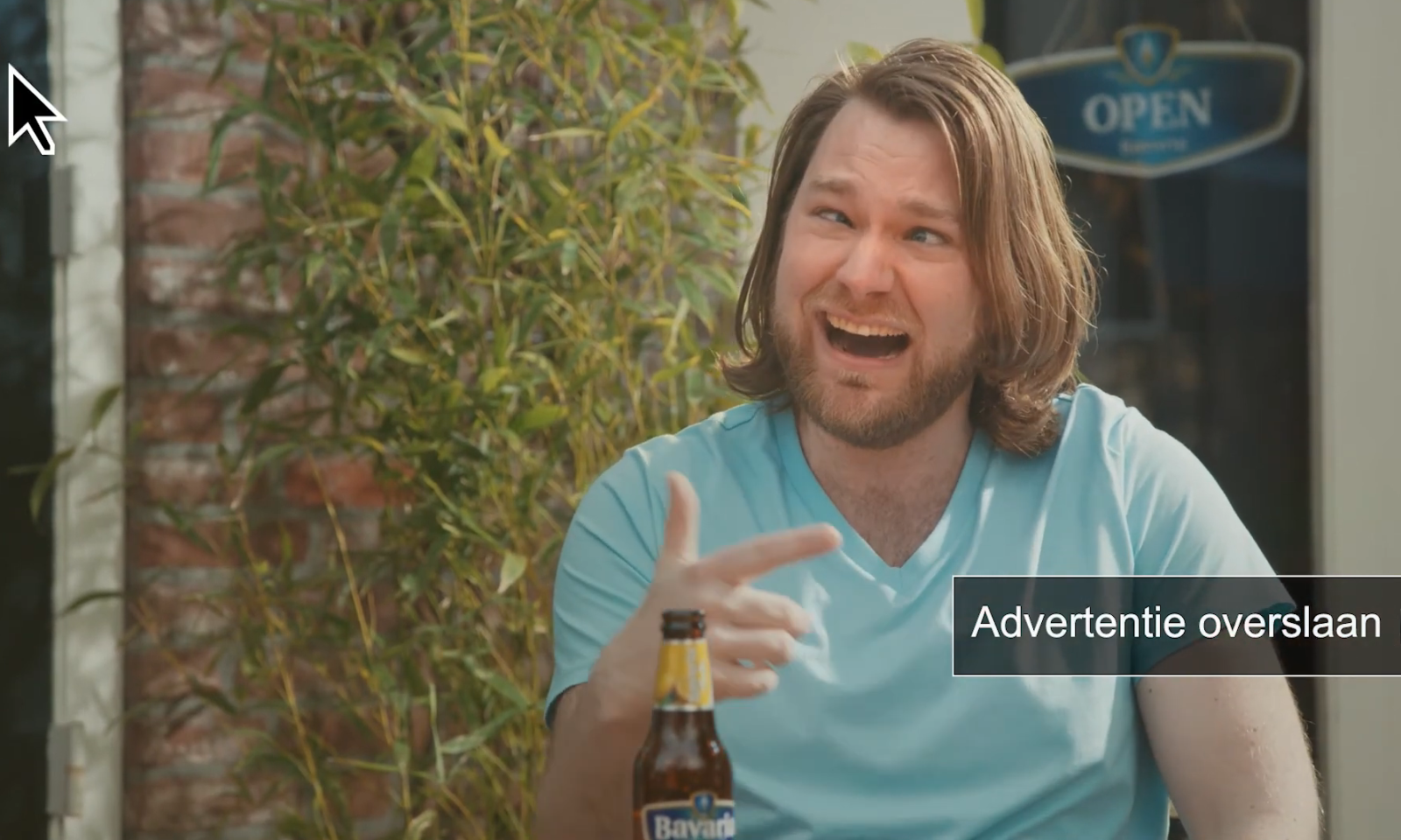 Man met halflang haar lacht en wijst, zittend aan tafel met Bavaria fles.