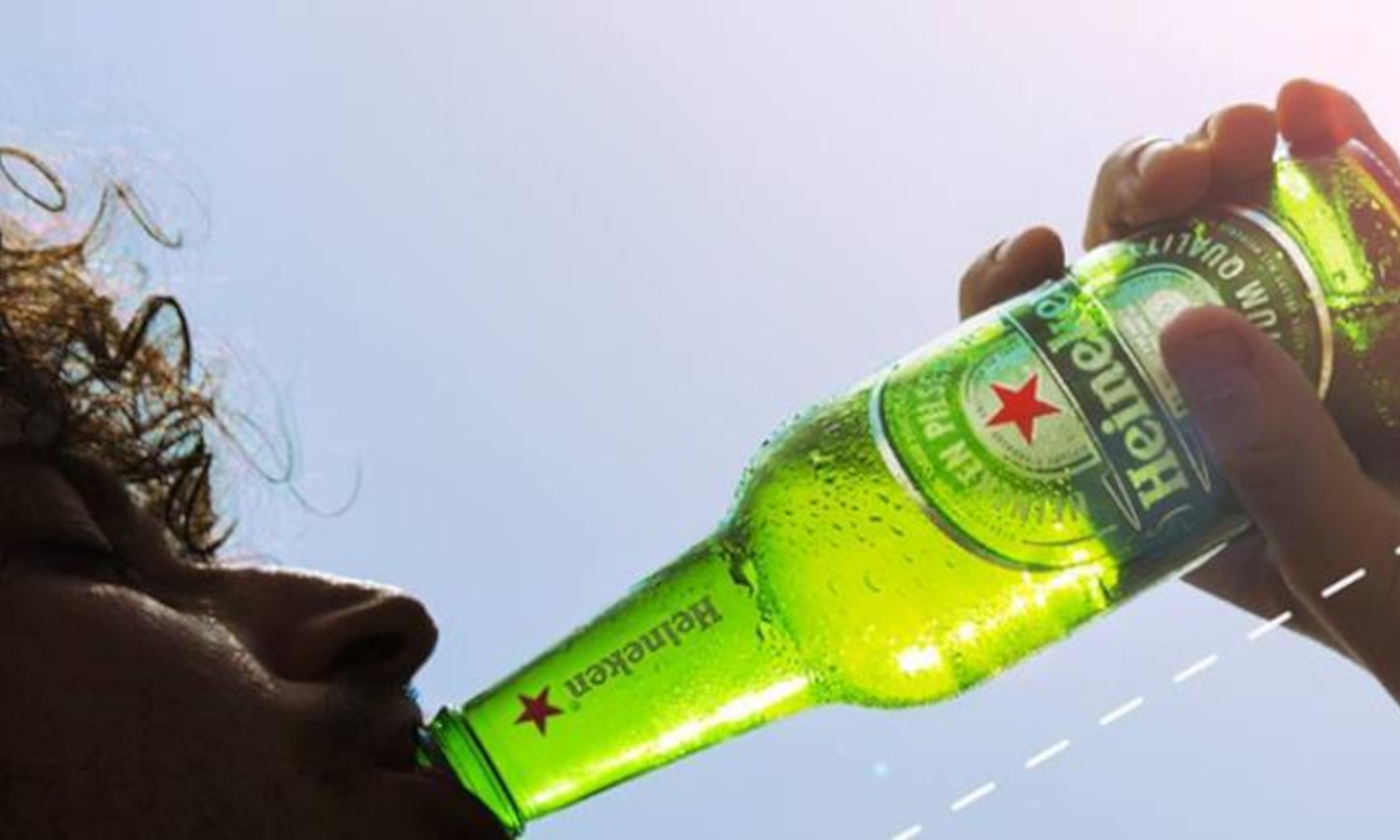 Close-up van een persoon die een groene Heineken fles drinkt bij zonnig weer.