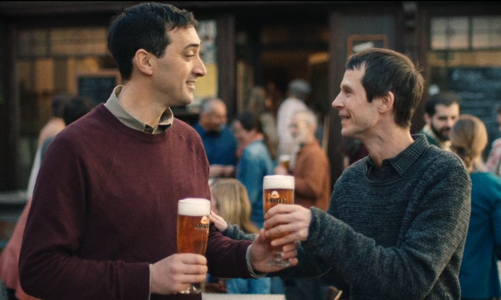 Twee mannen proosten met Amstel bier in een drukke buitenomgeving.