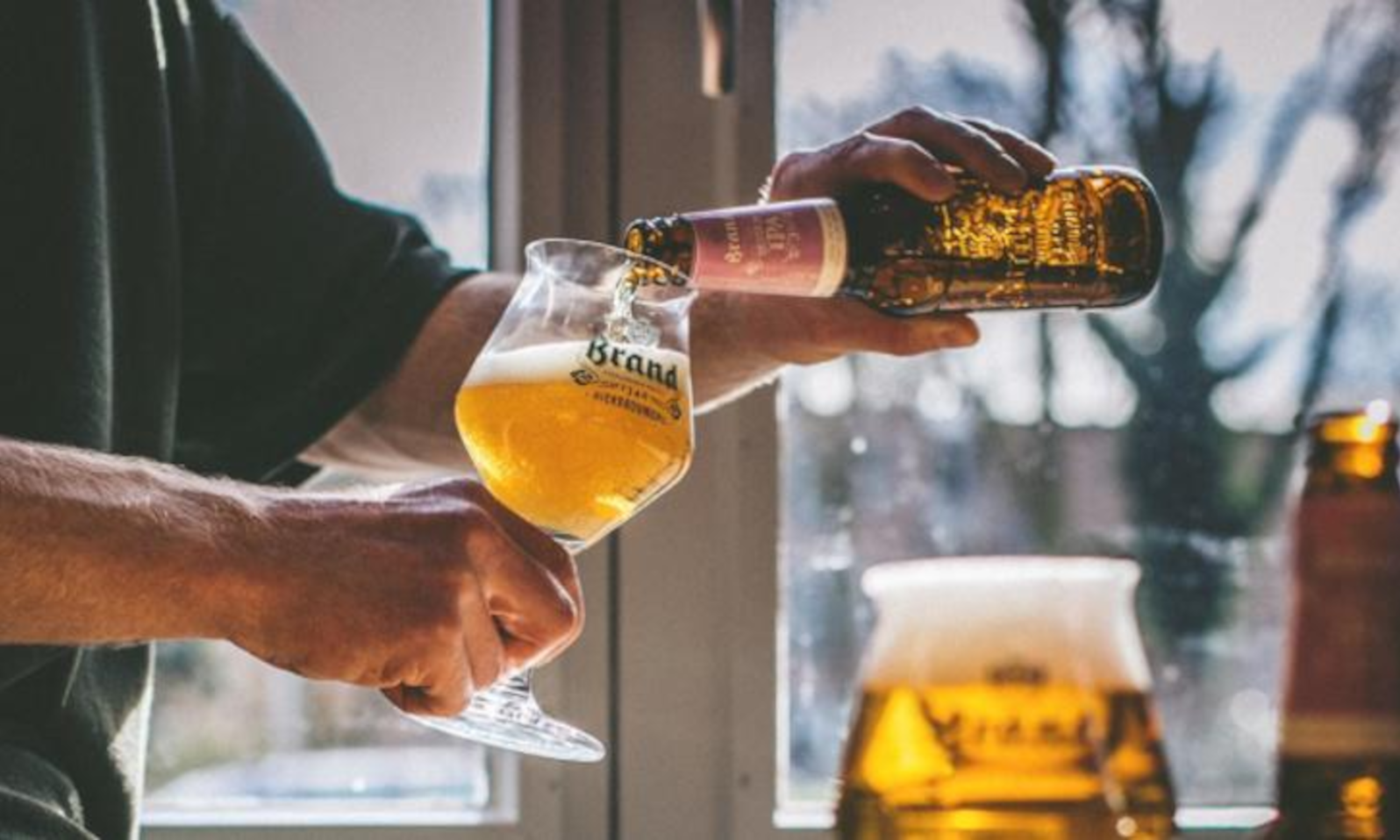 Iemand schenkt helder goudgeel bier in een Brand glas bij een raam.