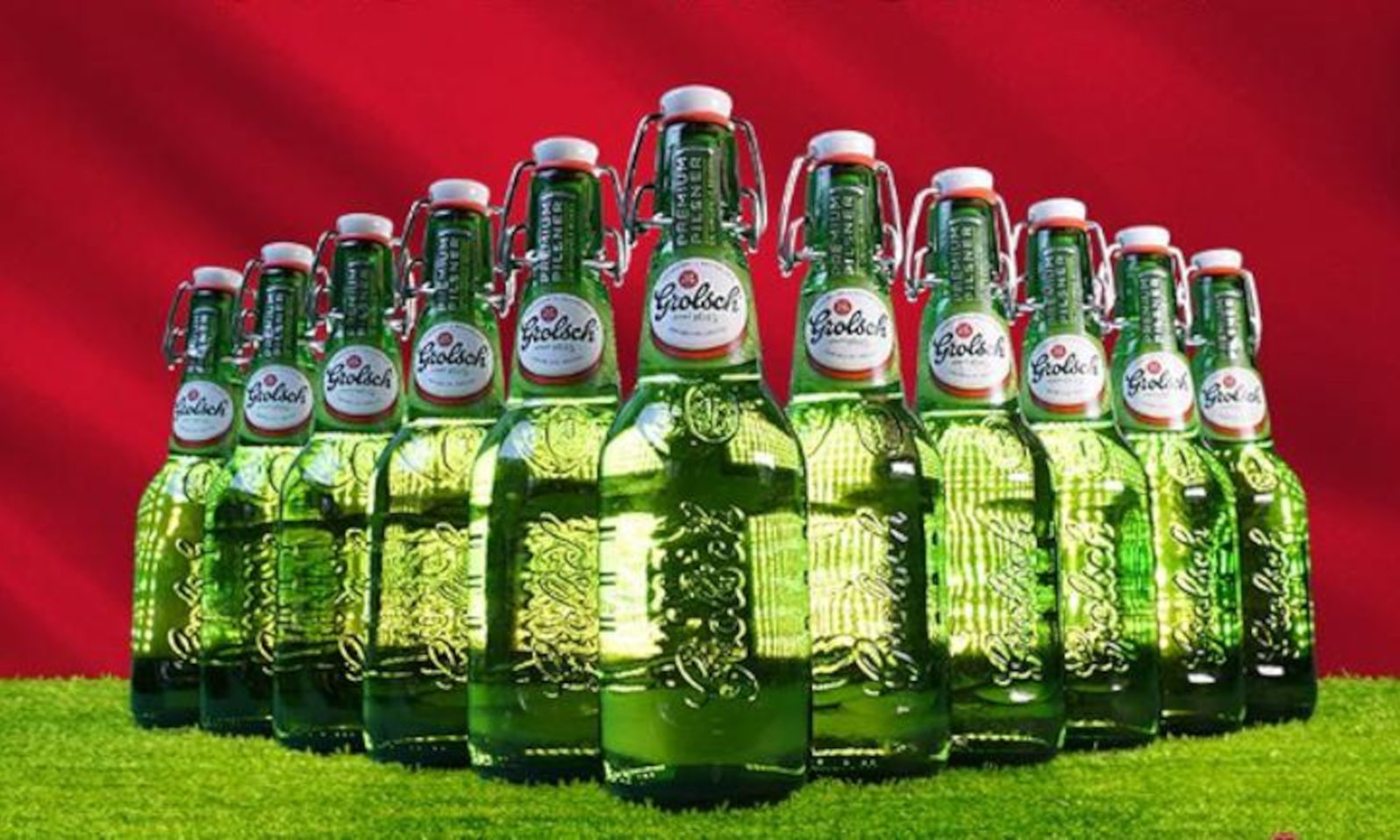 Elf groene Grolsch bierflessen in formatie op gras met rode achtergrond.