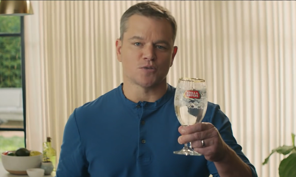 Man in blauw shirt houdt een leeg Stella Artois glas vast in een zonnige woonkamer.