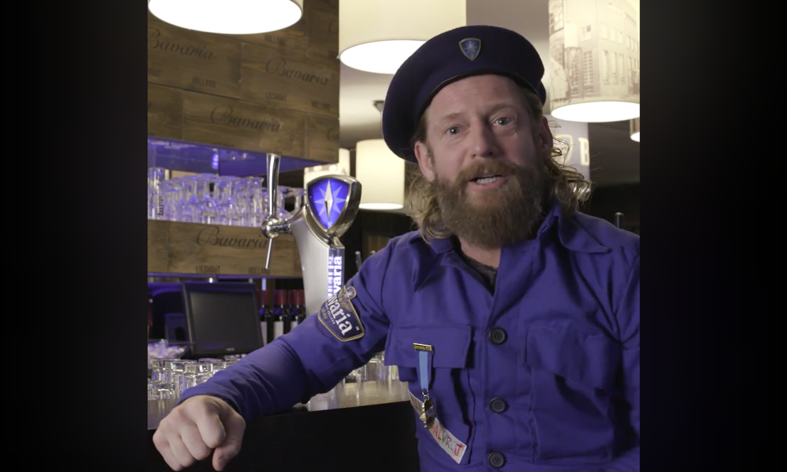 Man in blauw carnavalskostuum in een bar met Bavaria op de achtergrond.