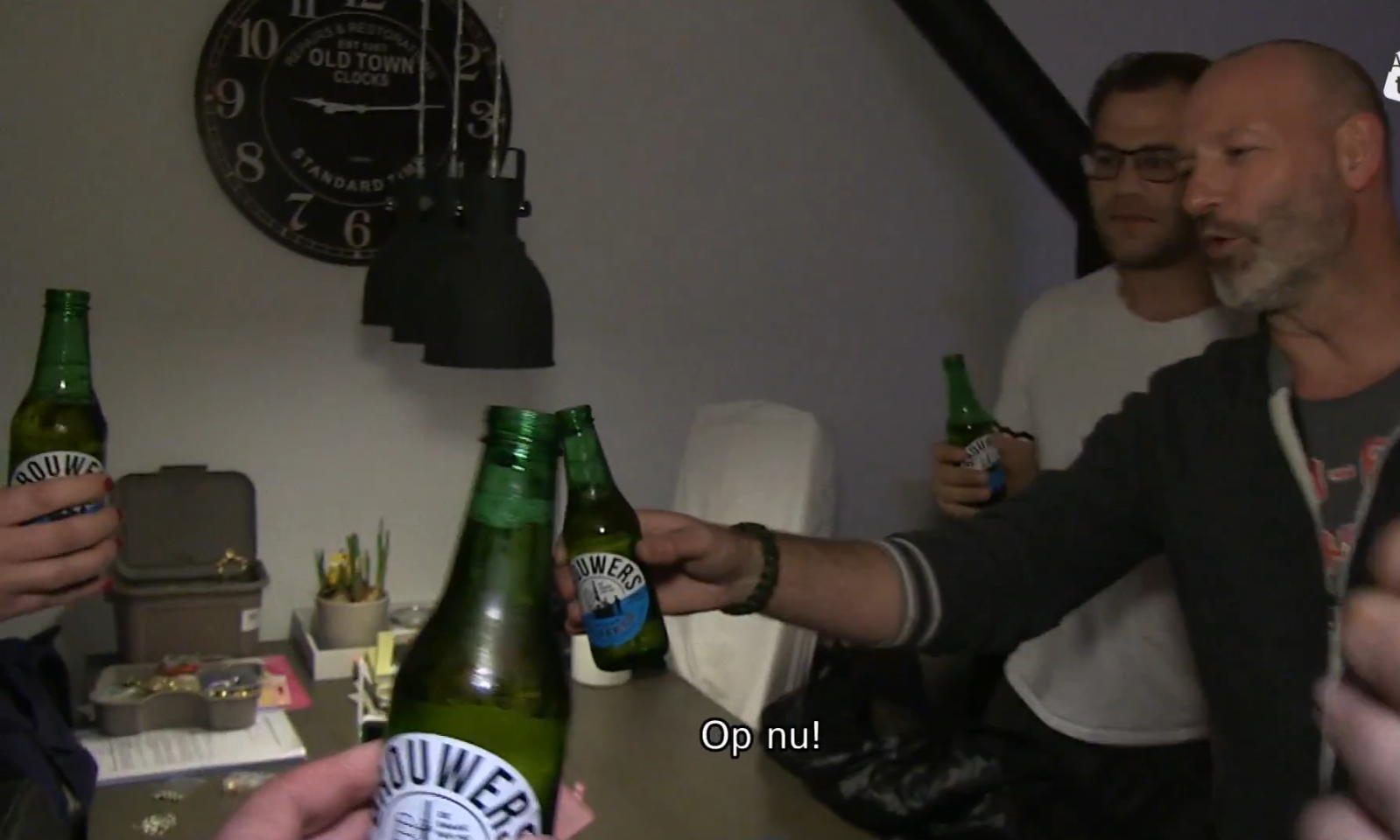 Groep mensen proost met groene bierflessen in een woonkamer met klok aan de muur.