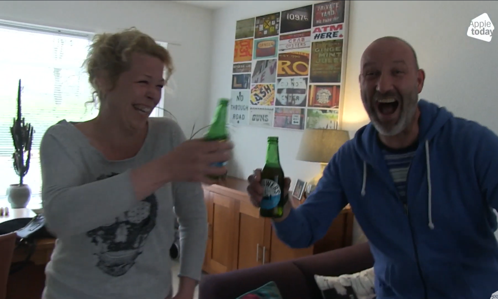 Twee lachende mensen die proosten met flesjes drank in een woonkamer.