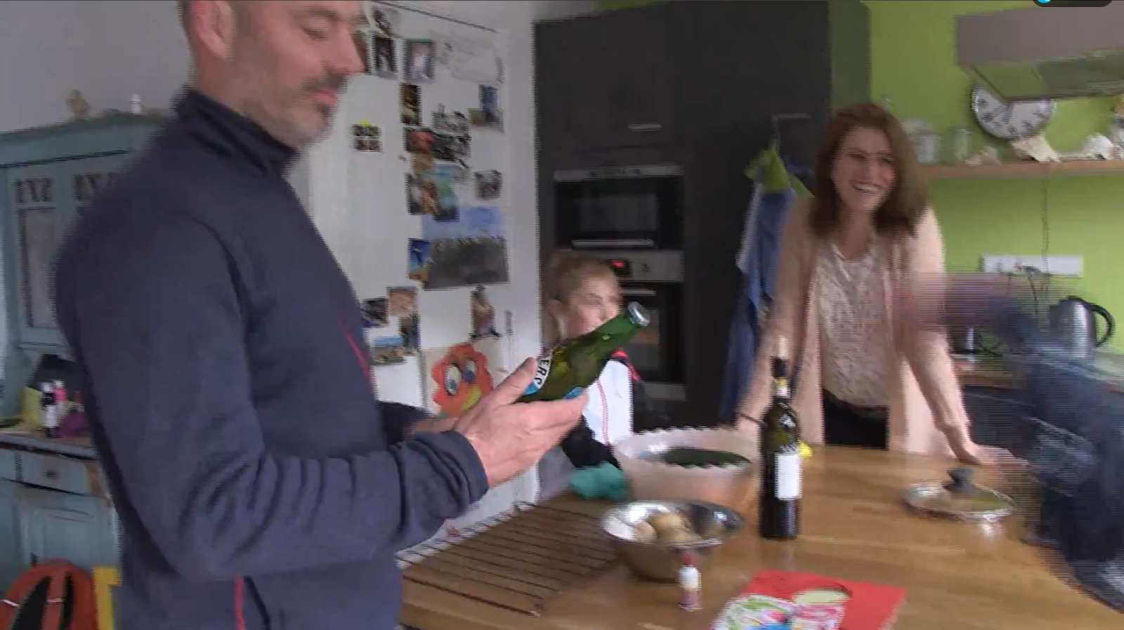 Gezin in keuken met man die fles inspecteert en vrouw die lacht naast een kind.