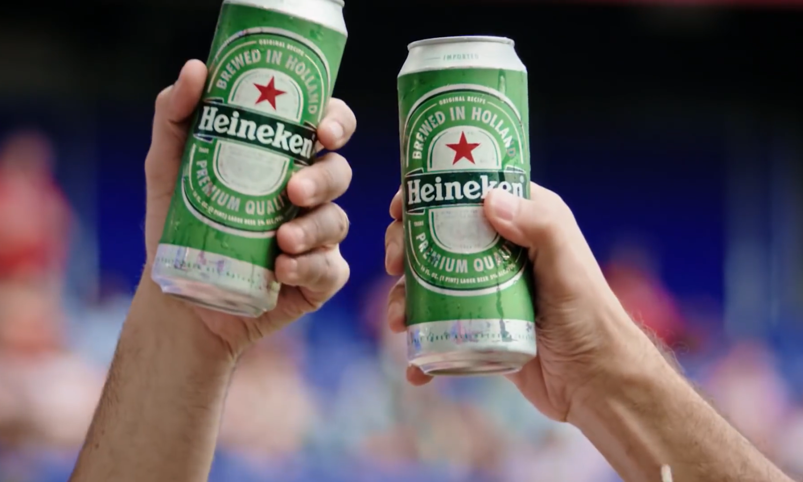 Twee handen houden Heineken-bierblikken op in een stadionachtige omgeving.
